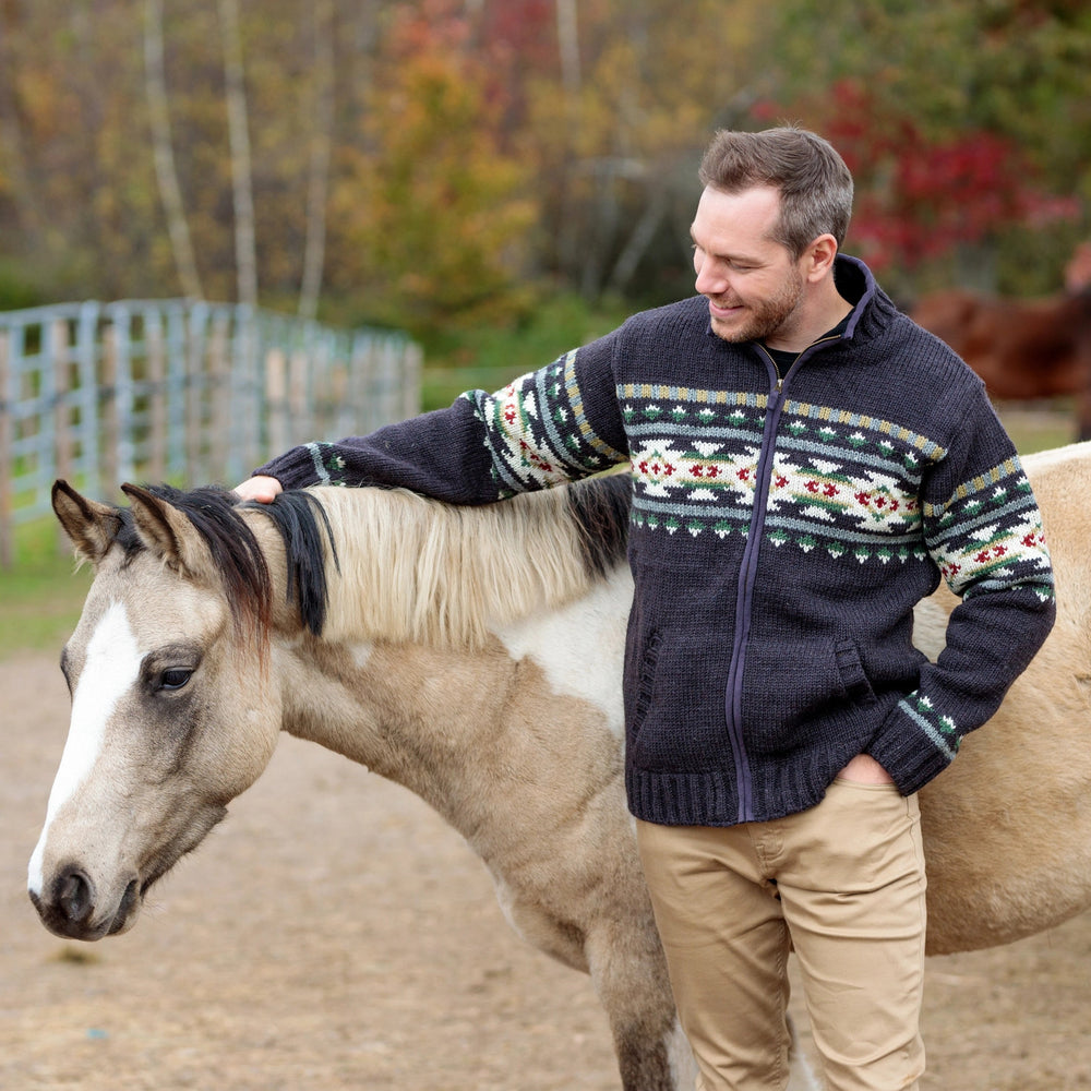 A man wearing a dark navy knit zip-up sweater with a colorful geometric Fair Isle pattern across the chest and cuffs, standing next to a light brown horse.